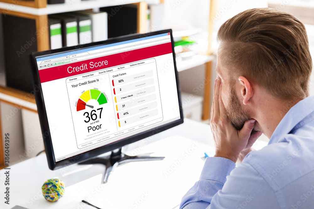 Person sitting at a desk viewing a credit score report on a computer monitor. The screen displays a score of 367 out of 900, labeled as 'Poor', with a color-coded gauge and breakdown of credit factors including payment history, credit utilization, and types of credit used