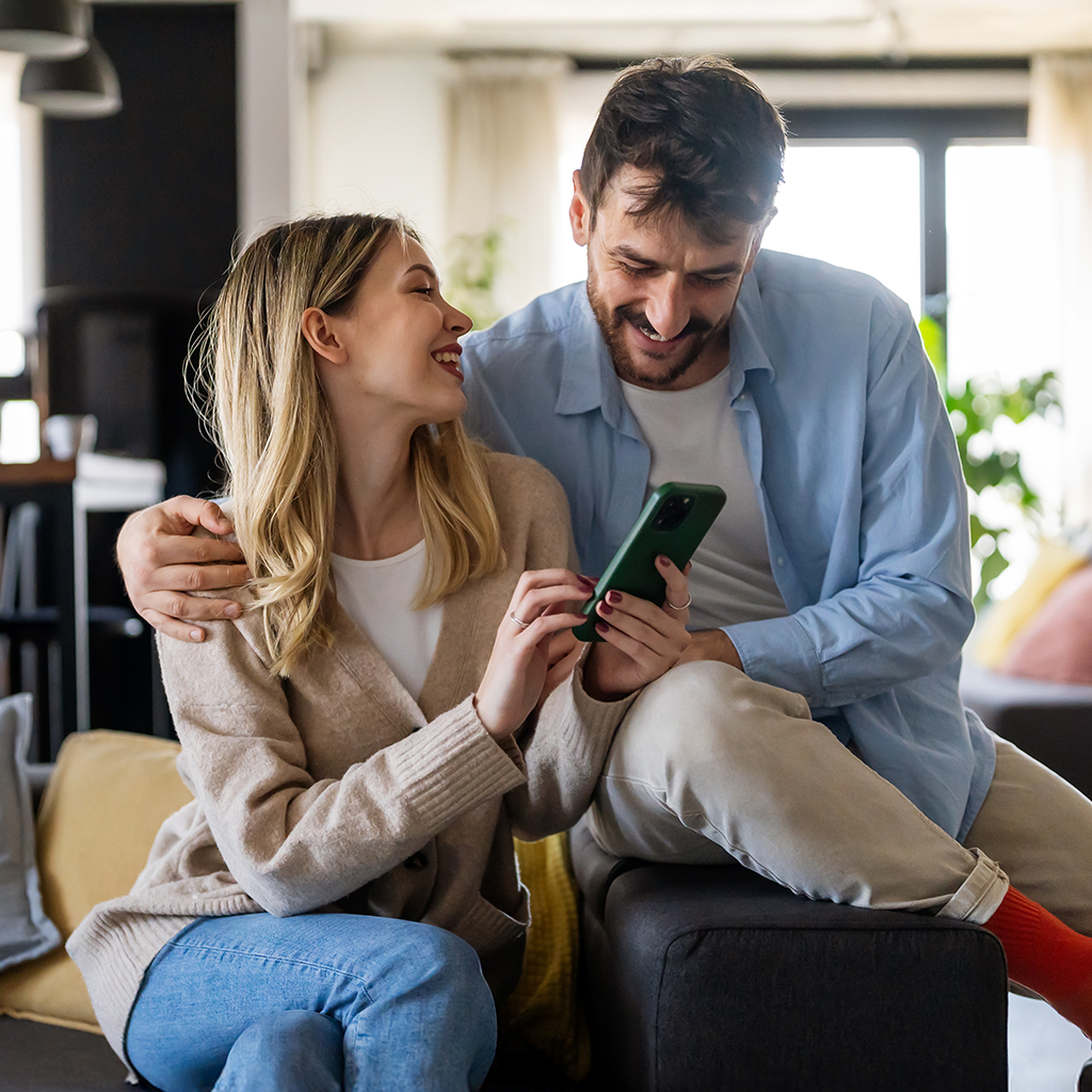 Two people sitting closely on a couch, one holding a smartphone, with natural light coming through windows