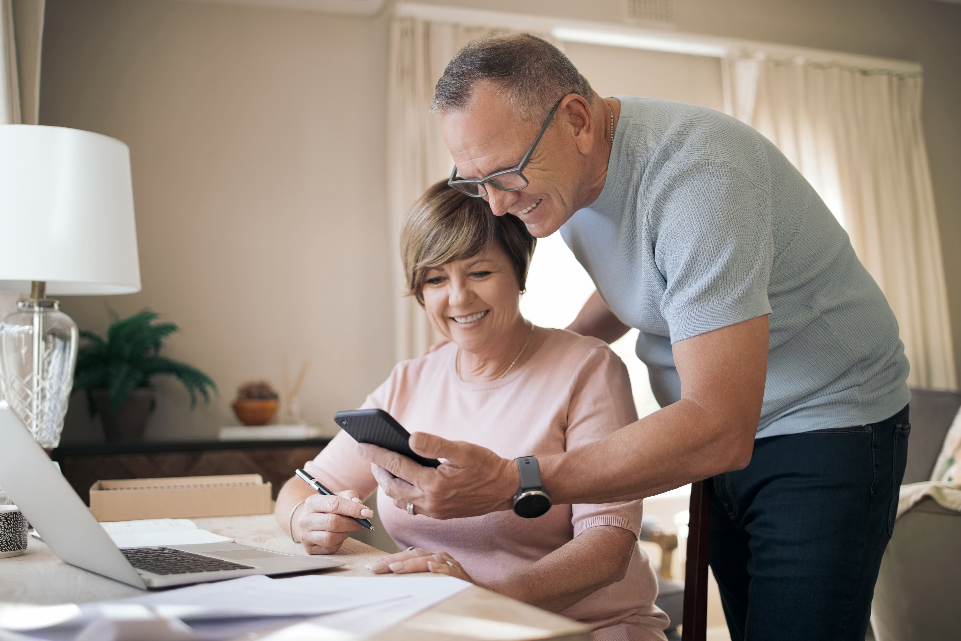 Two people in home setting, one at desk with laptop and papers, other showing smartphone