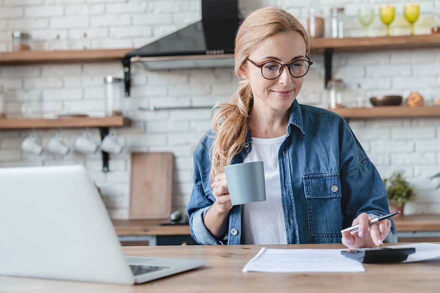 Woman smiling when standing in the kitchen with cup in her hand and calculator in front of her, on a desk