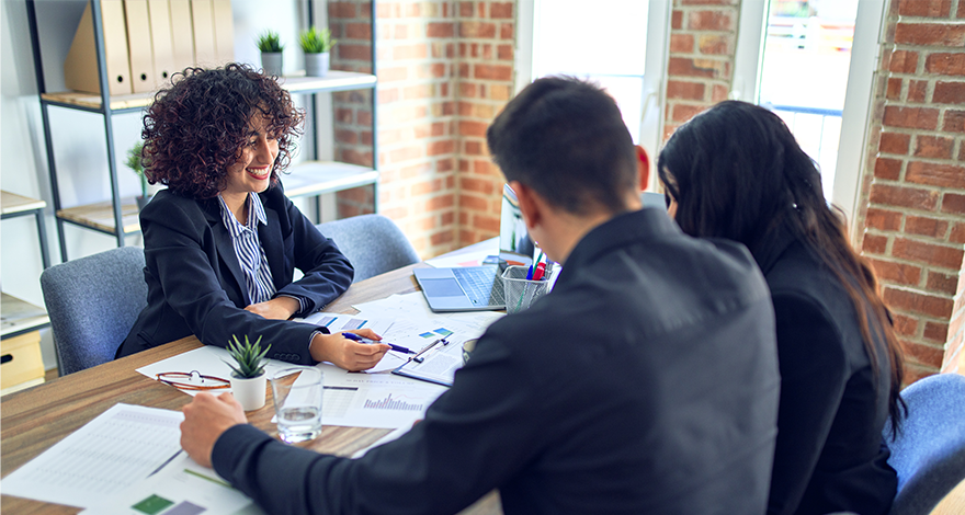 Three people in an office setting sitting around a table covered with financial documents, charts, and a laptop